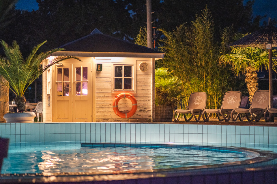 Outdoorschwimmbecken mit Holzhütte dahinter und Sonnenliegen bei Nacht beleuchtet