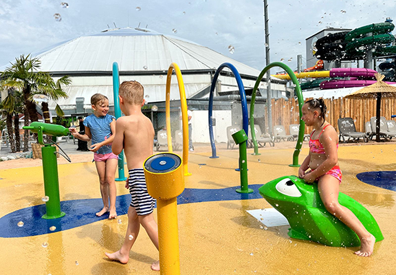Drei Kinder in Badeklamotten die auf einem Wasserspielplatz spielen im Hintergrund bunte Rutschen